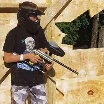 Person in paintball gear holding a gun, inside a wooden structure.
