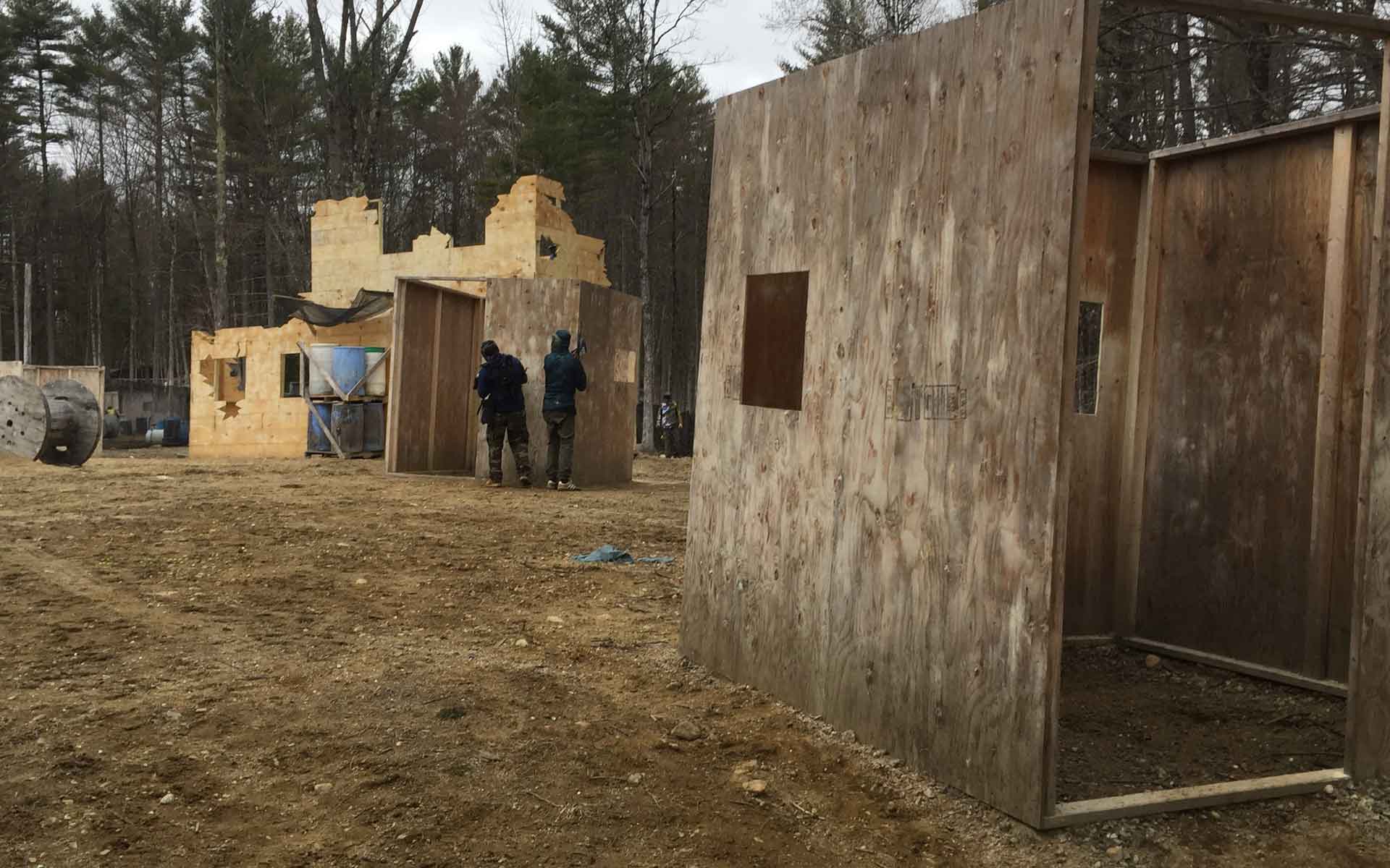 Paintball field with players near wooden structures and a ruined building.