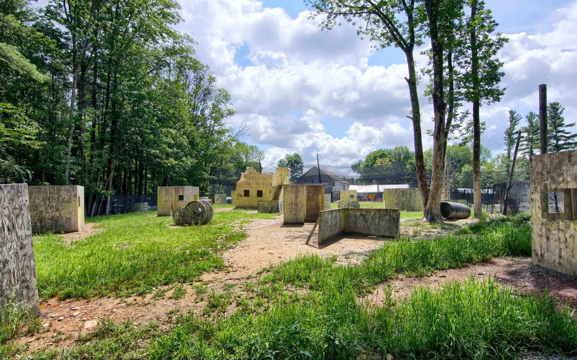 Paintball field with bunkers and buildings amidst trees and grass under a cloudy sky.