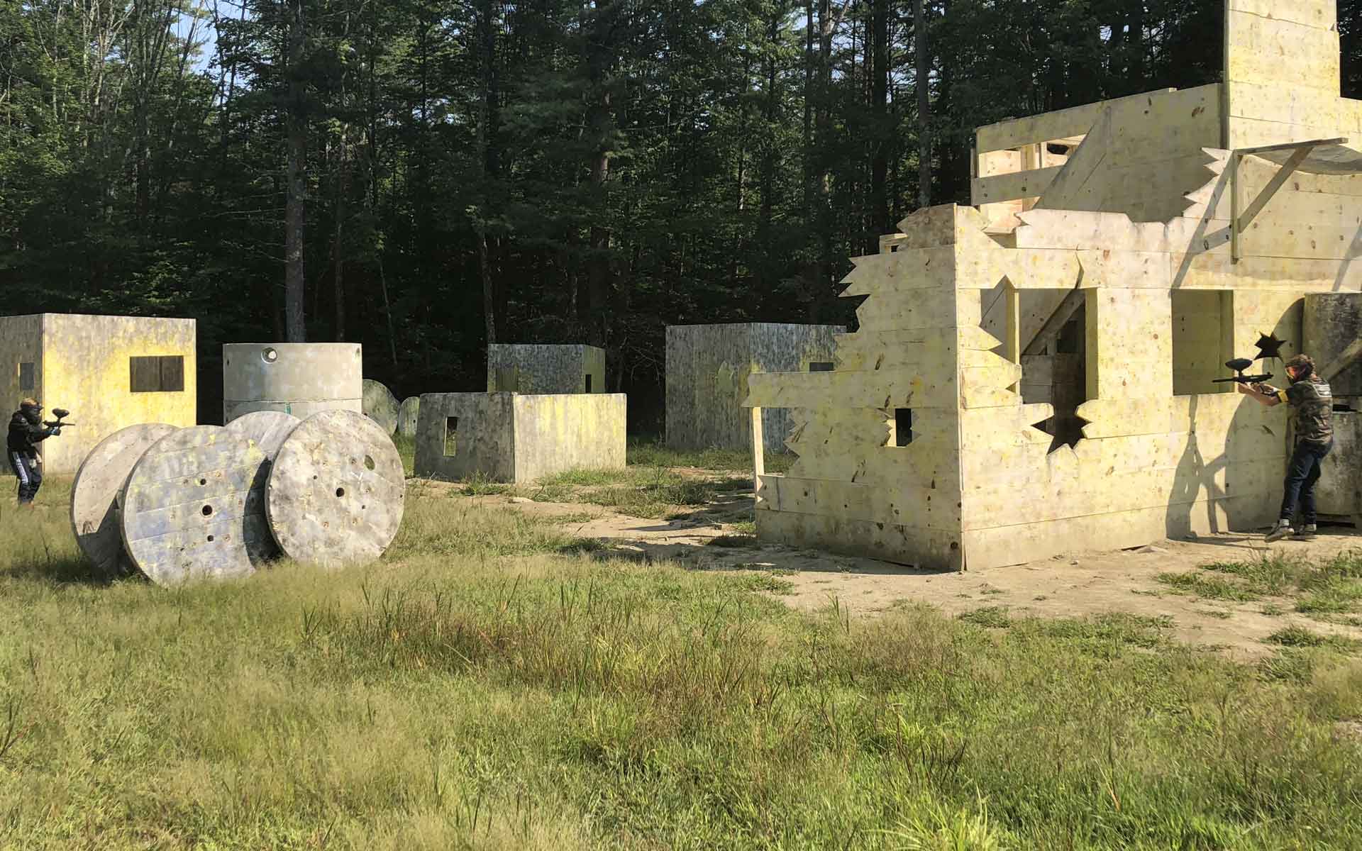 Paintball game in progress: Players shooting at each other near bunkers and dilapidated structures outdoors.