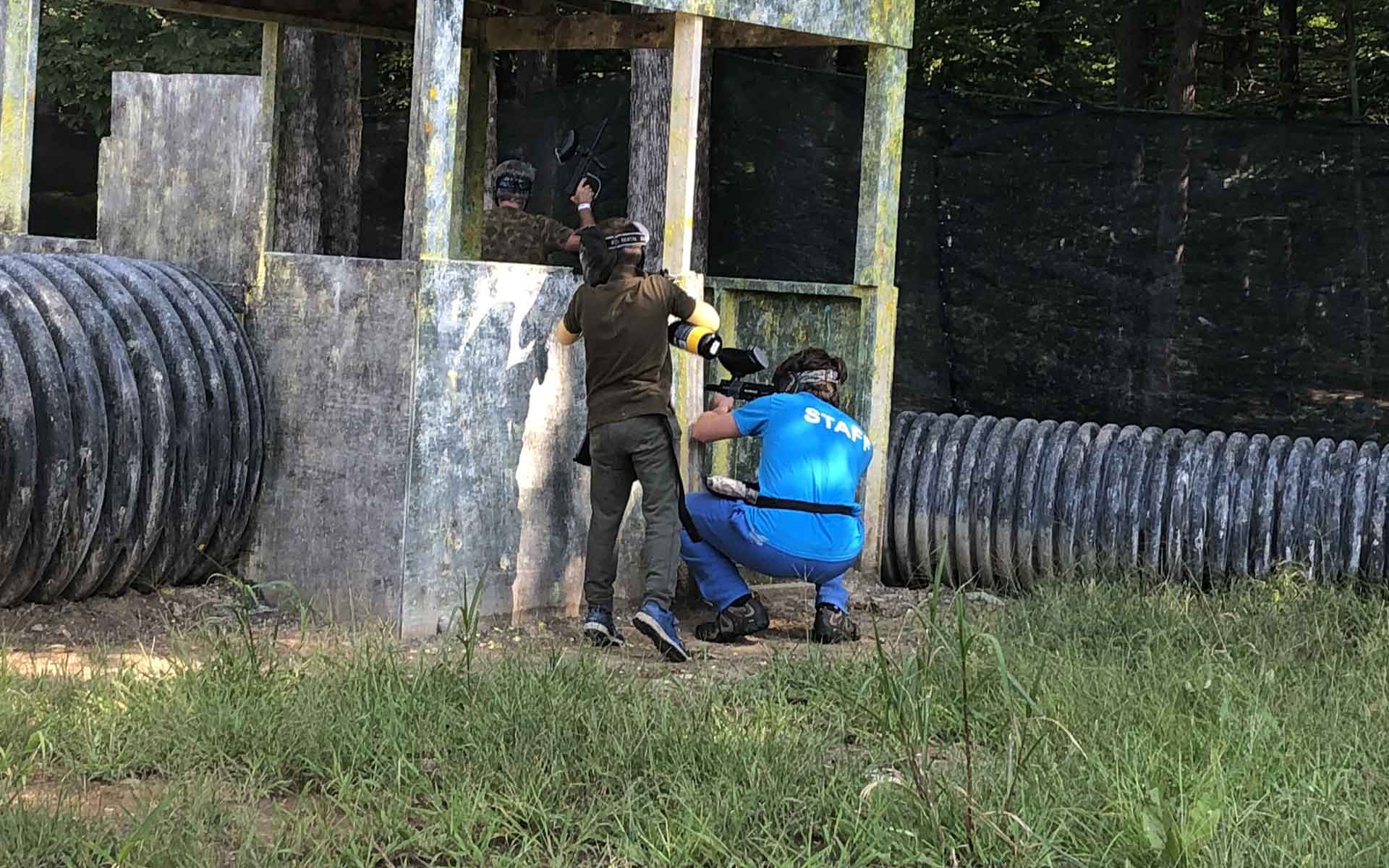 People playing paintball, outdoors; person aiming gun from behind bunker, another crouched with a gun.