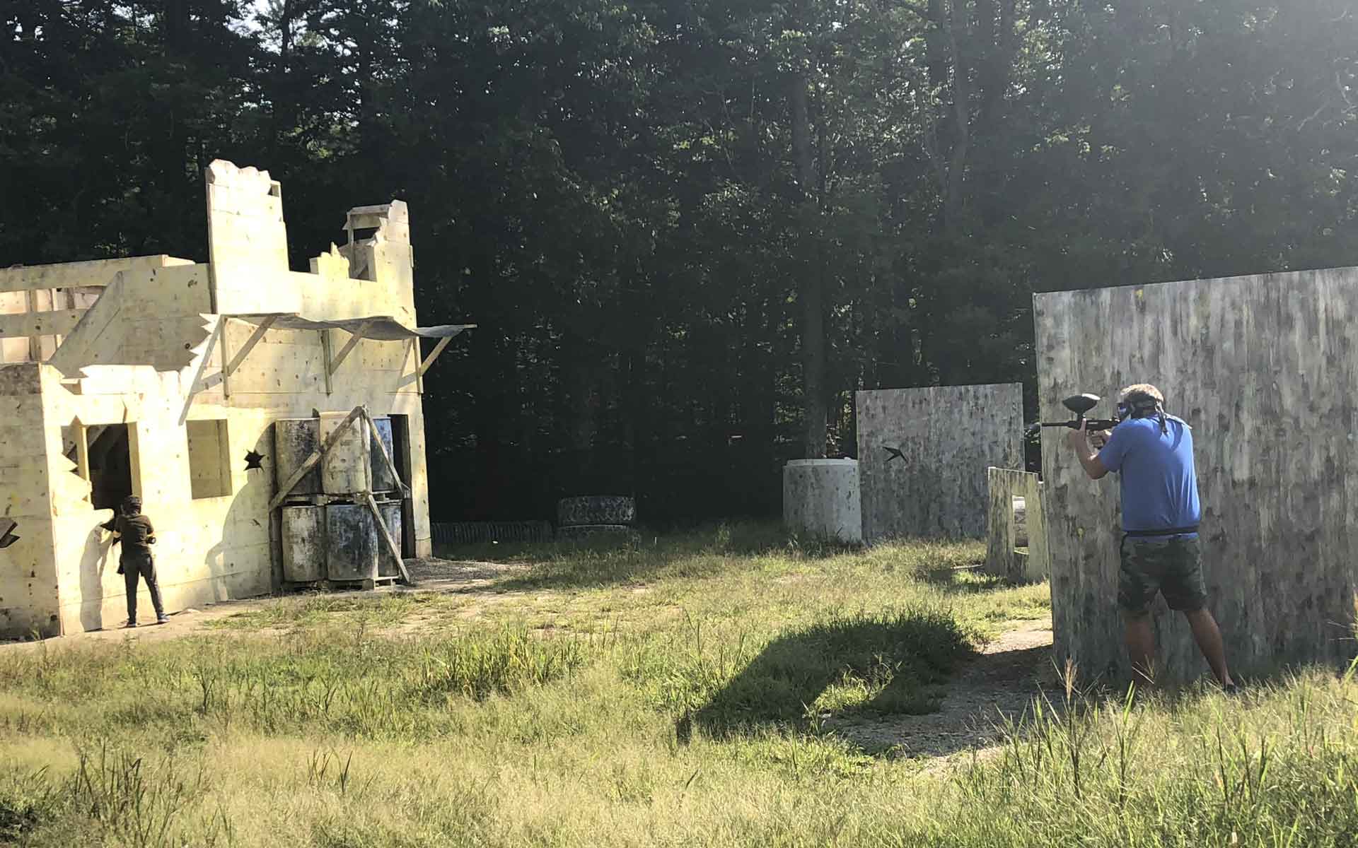 Two people playing paintball in a wooded area. One shoots from behind a wooden wall, another aims from a building.