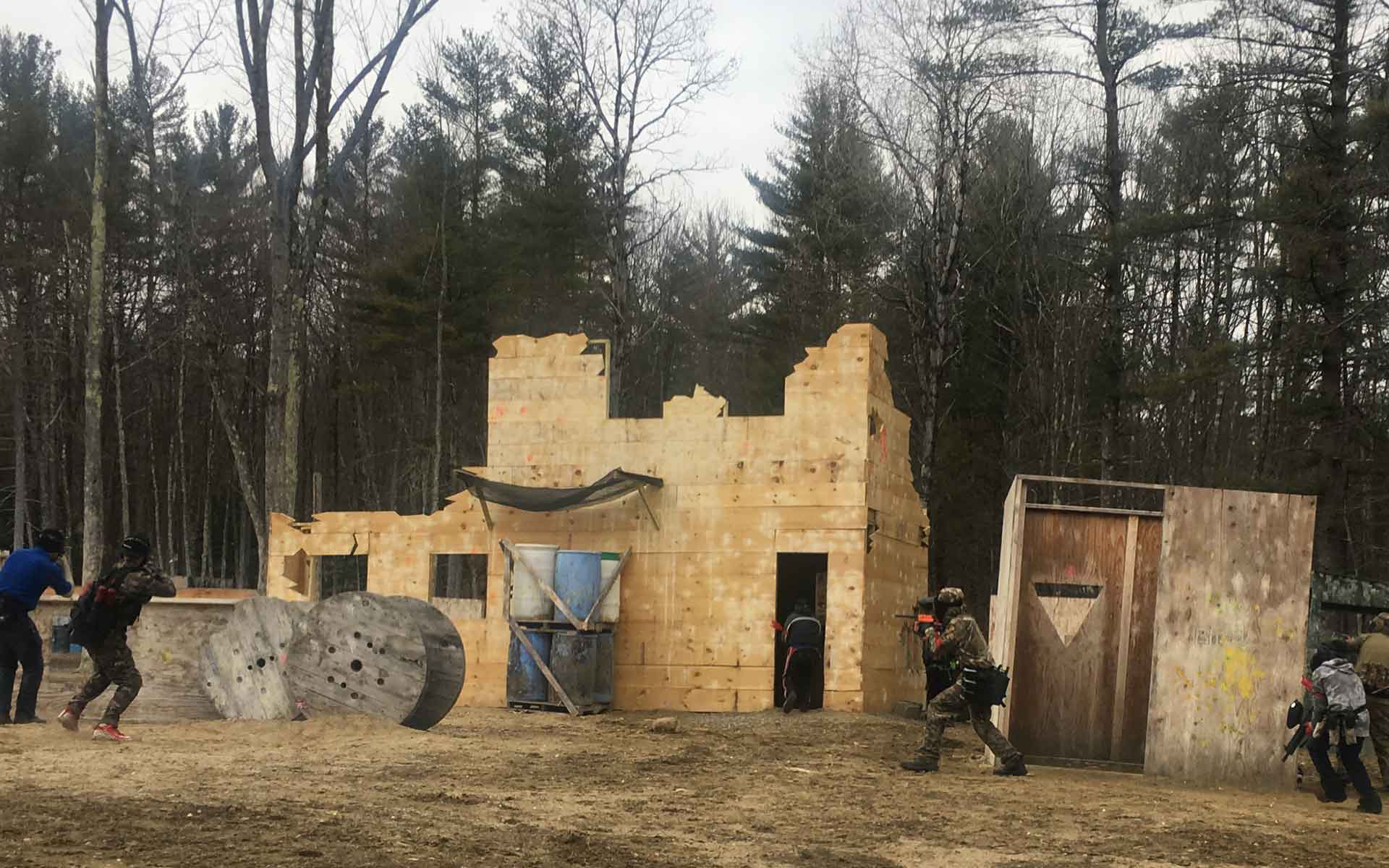 Paintball players in action at an outdoor playing field. Structures and forest in background.