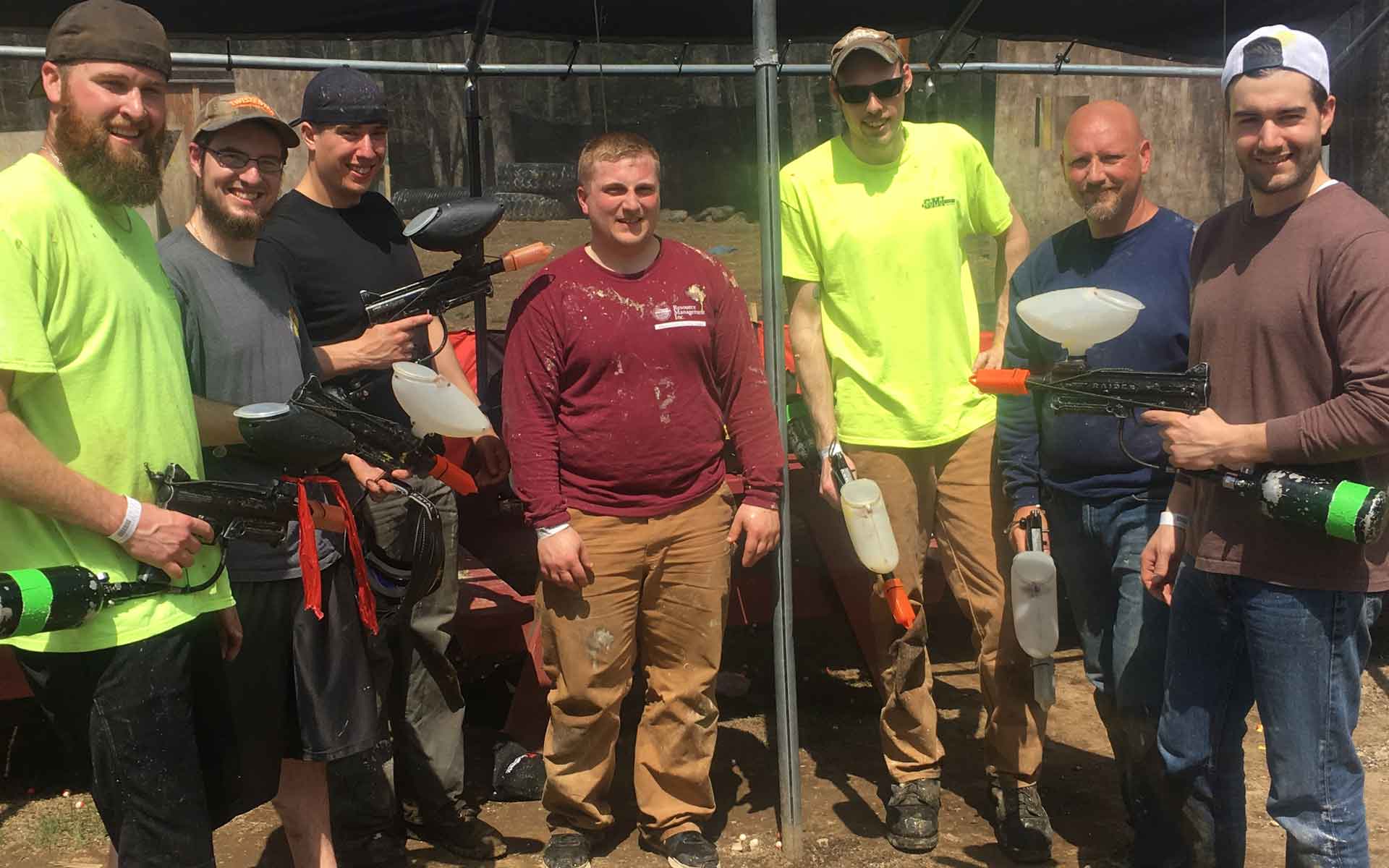 Group of men posing with paintball guns; outdoors, under shade.