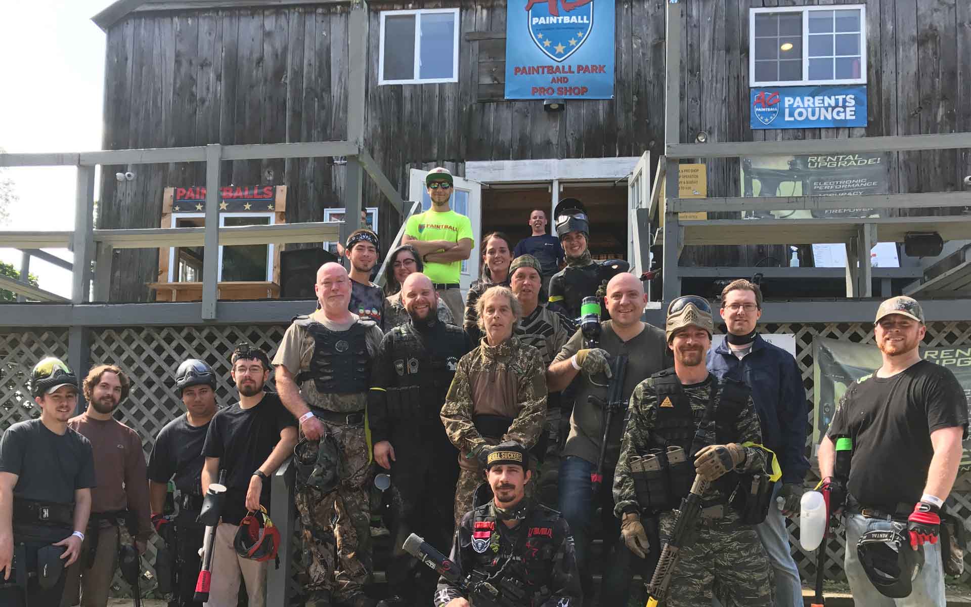 Group of people, some in camouflage, posing on wooden stairs of a building, likely a paintball venue.