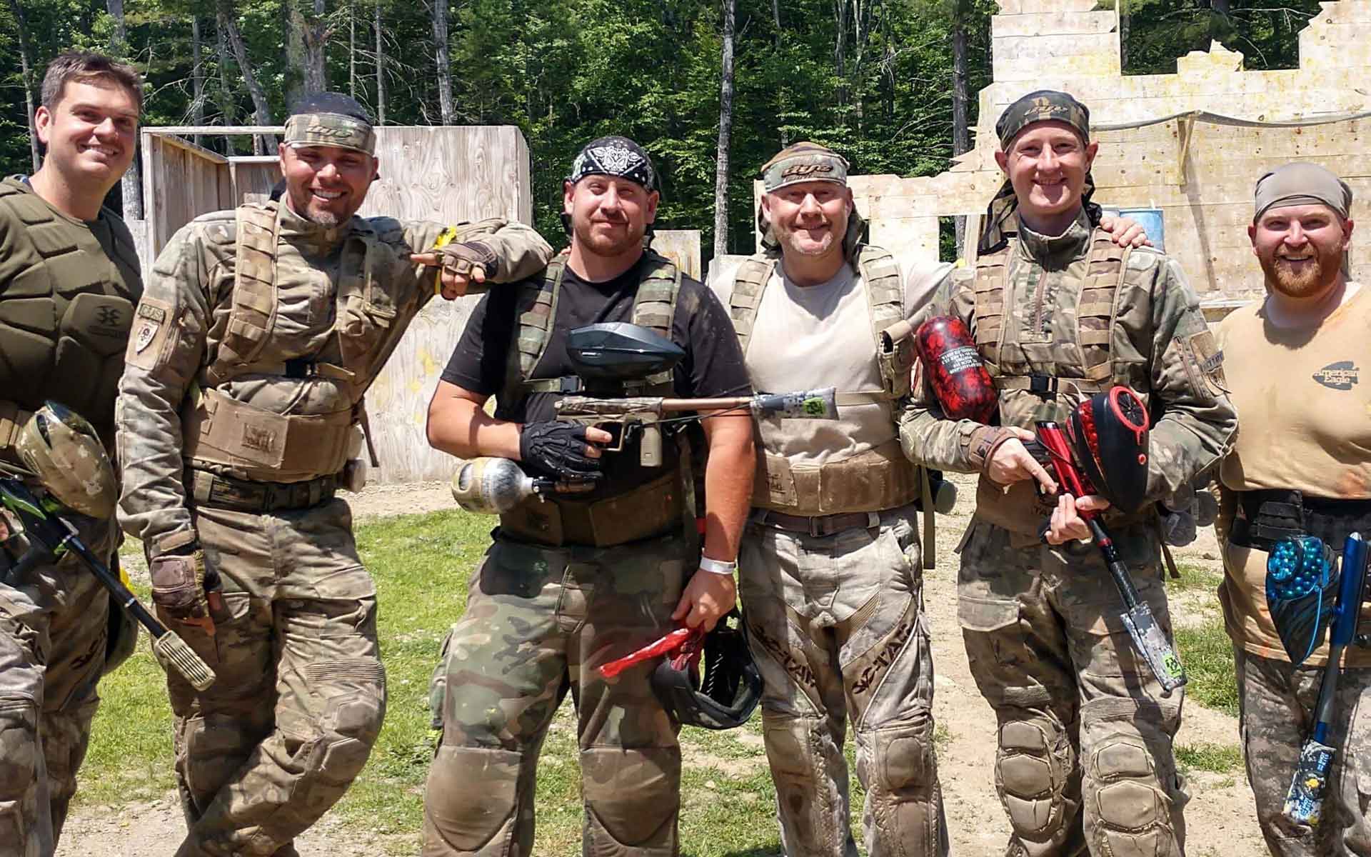Six men in camouflage gear pose with paintball guns on a paintball field.