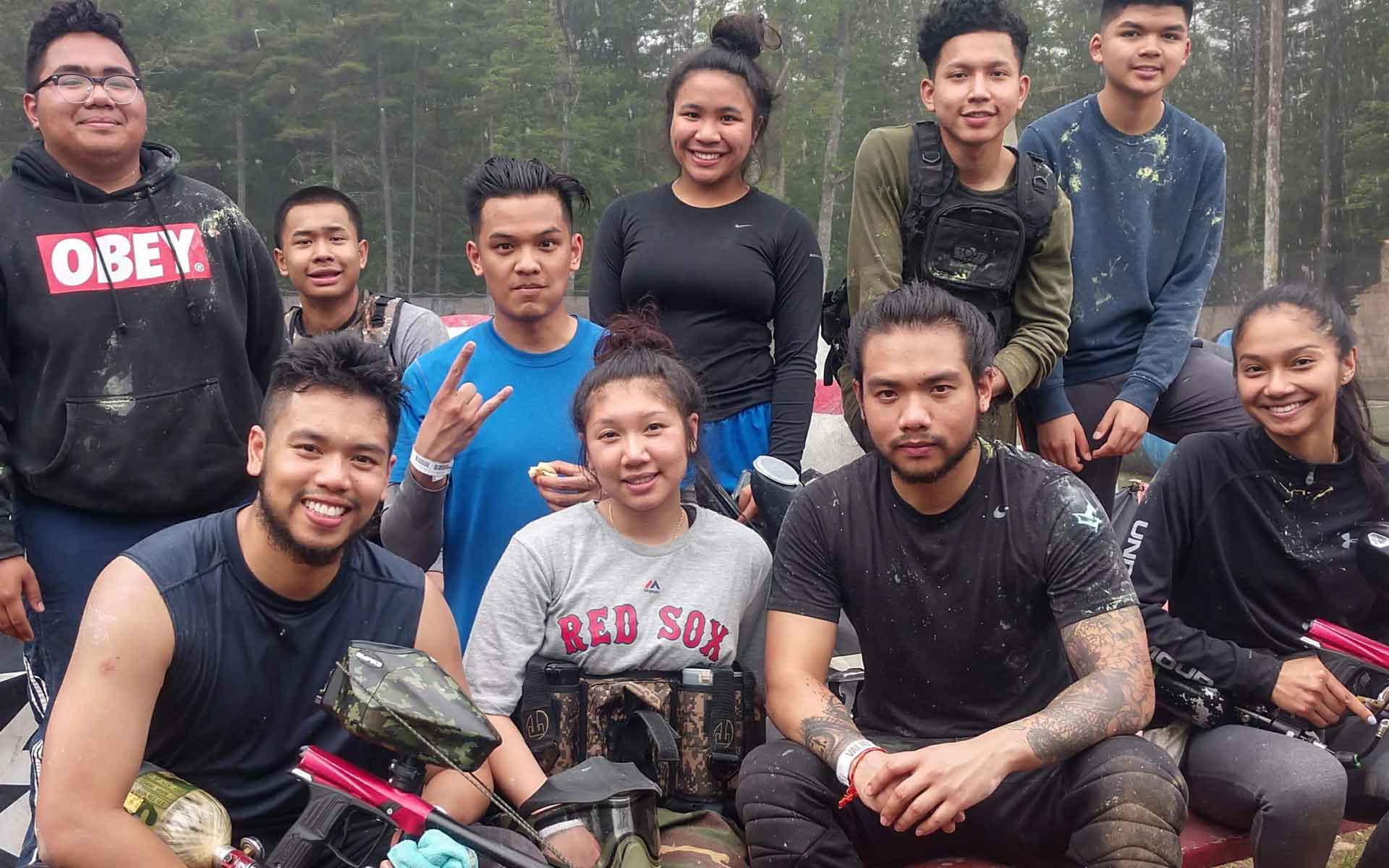 Group of diverse young adults, some holding paintball guns, smiling outdoors, possibly at a paintball arena.