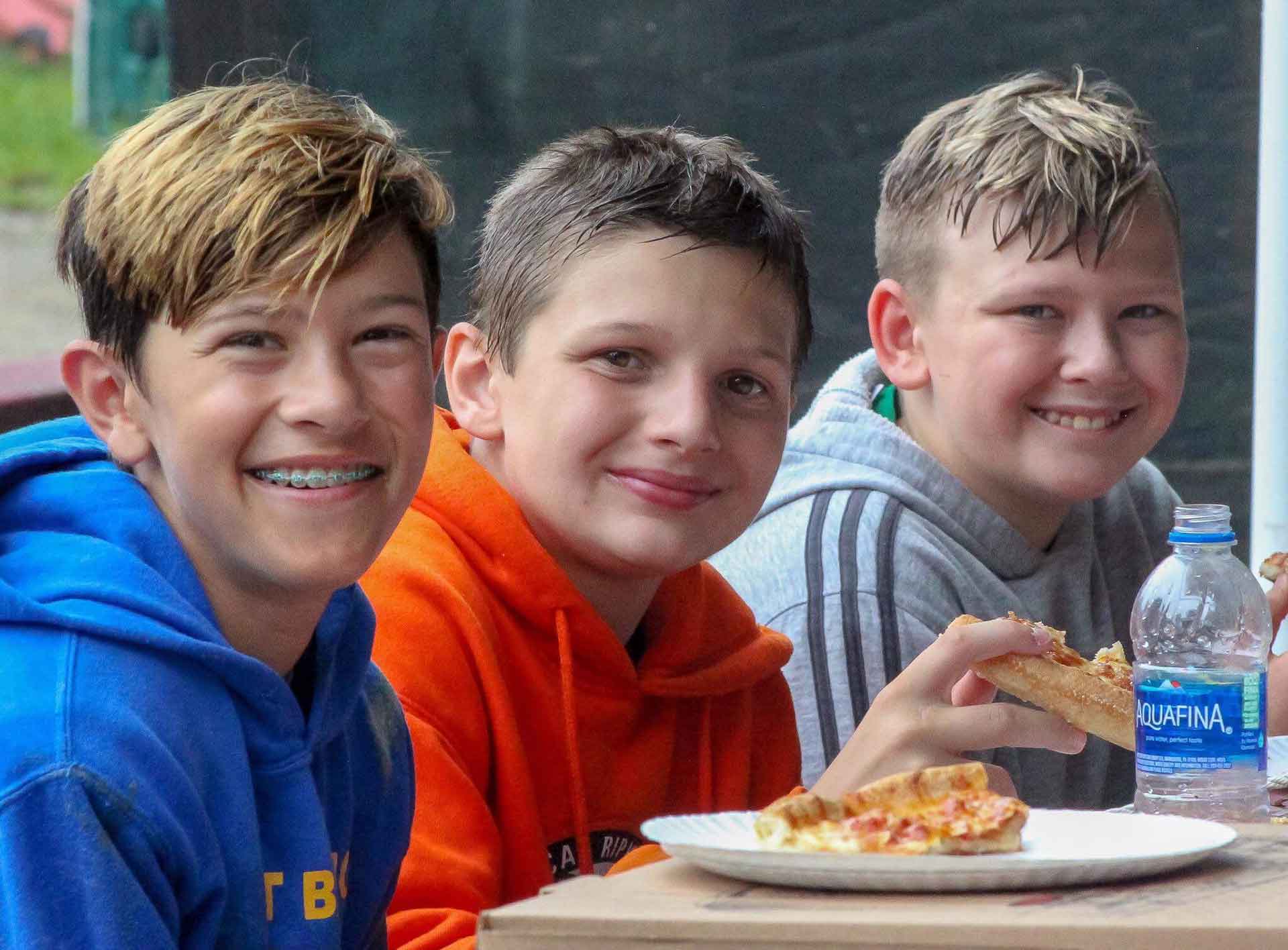 Three smiling boys eating pizza outdoors. One with braces in blue hoodie, others in orange and grey.
