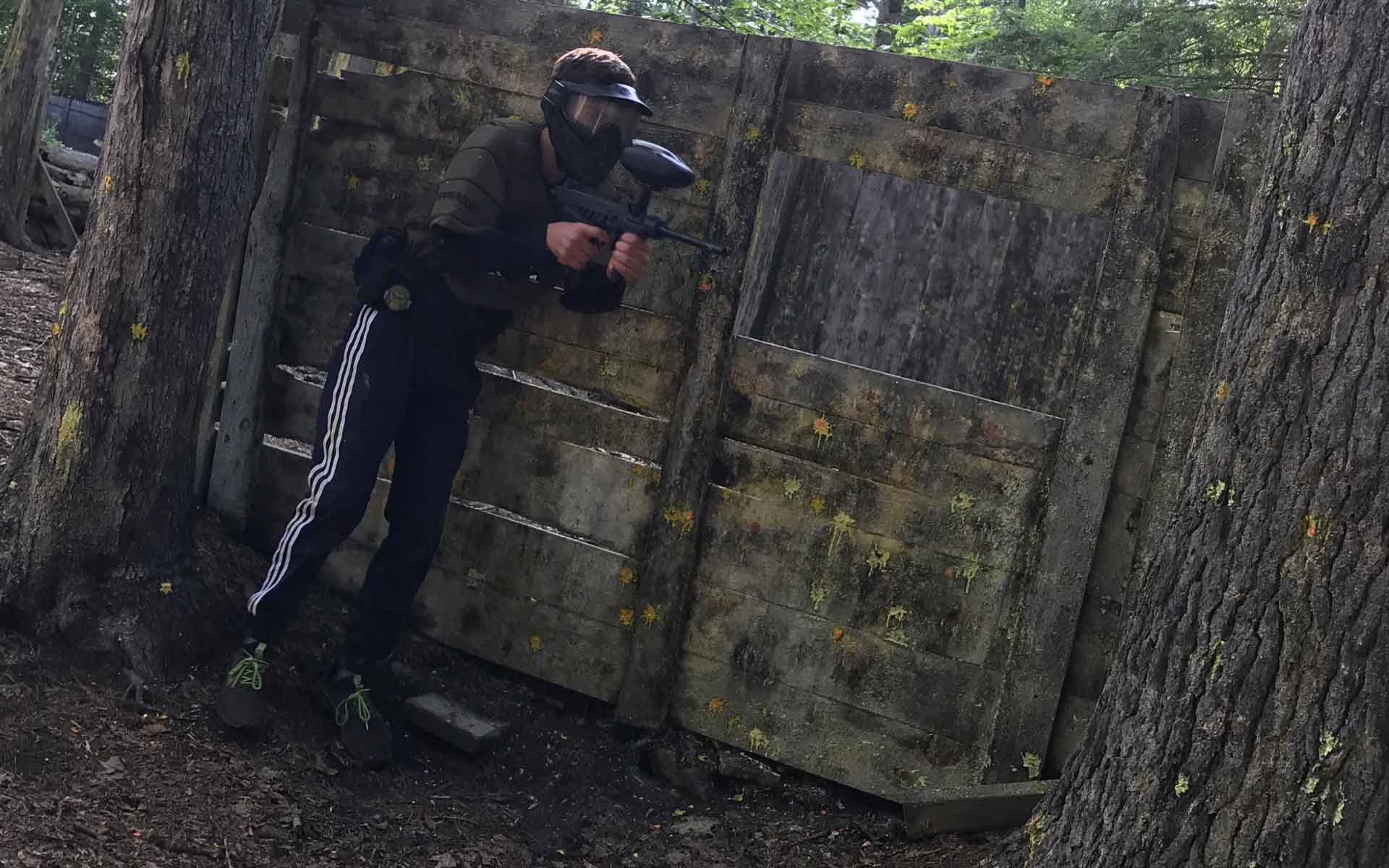 Person playing paintball, aiming a marker gun from behind a wooden barricade in a wooded area.