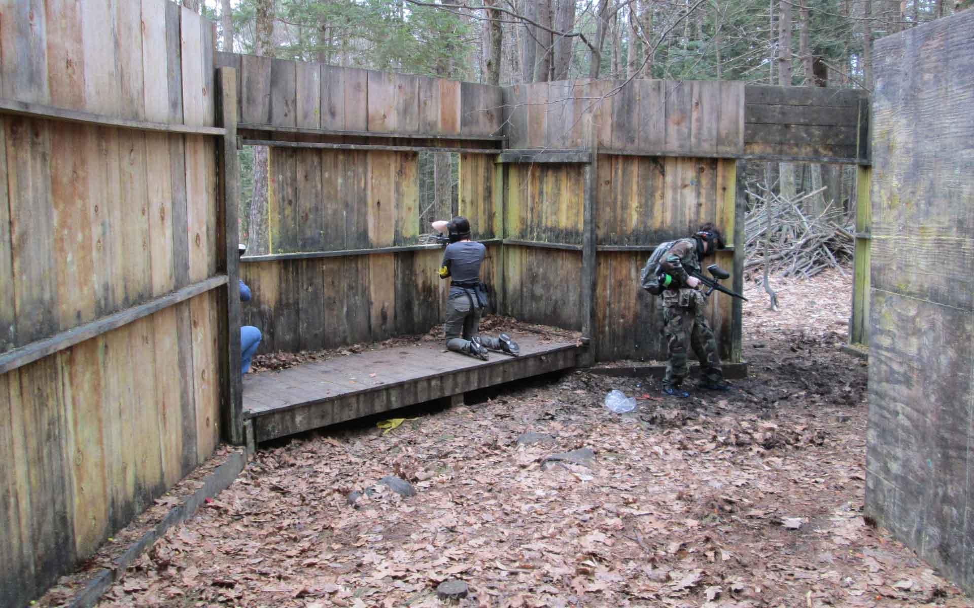 Paintball players in camouflage, aiming guns from wooden structures in a wooded area.