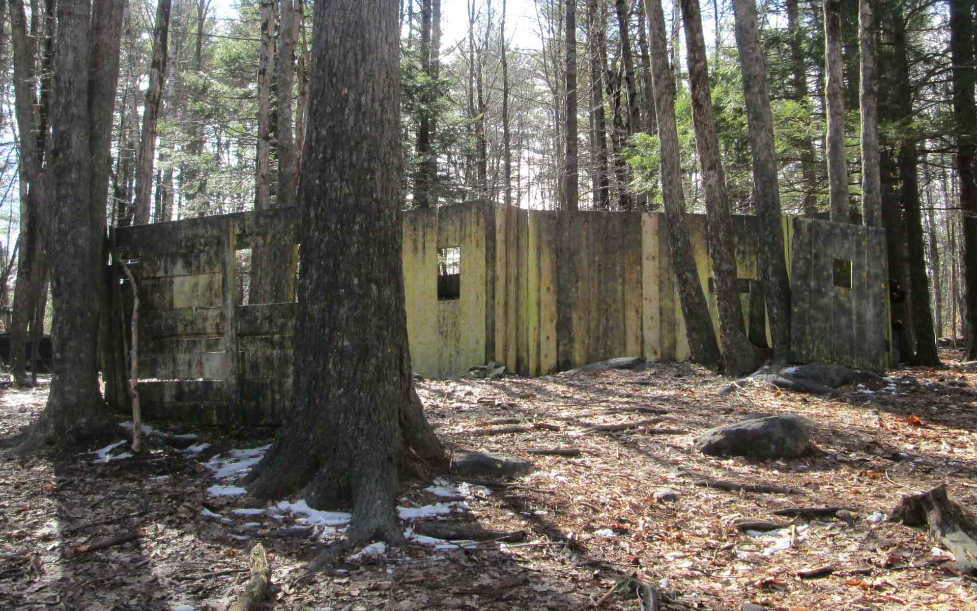 Old, abandoned concrete building in a forest, surrounded by trees. Some snow on the ground.