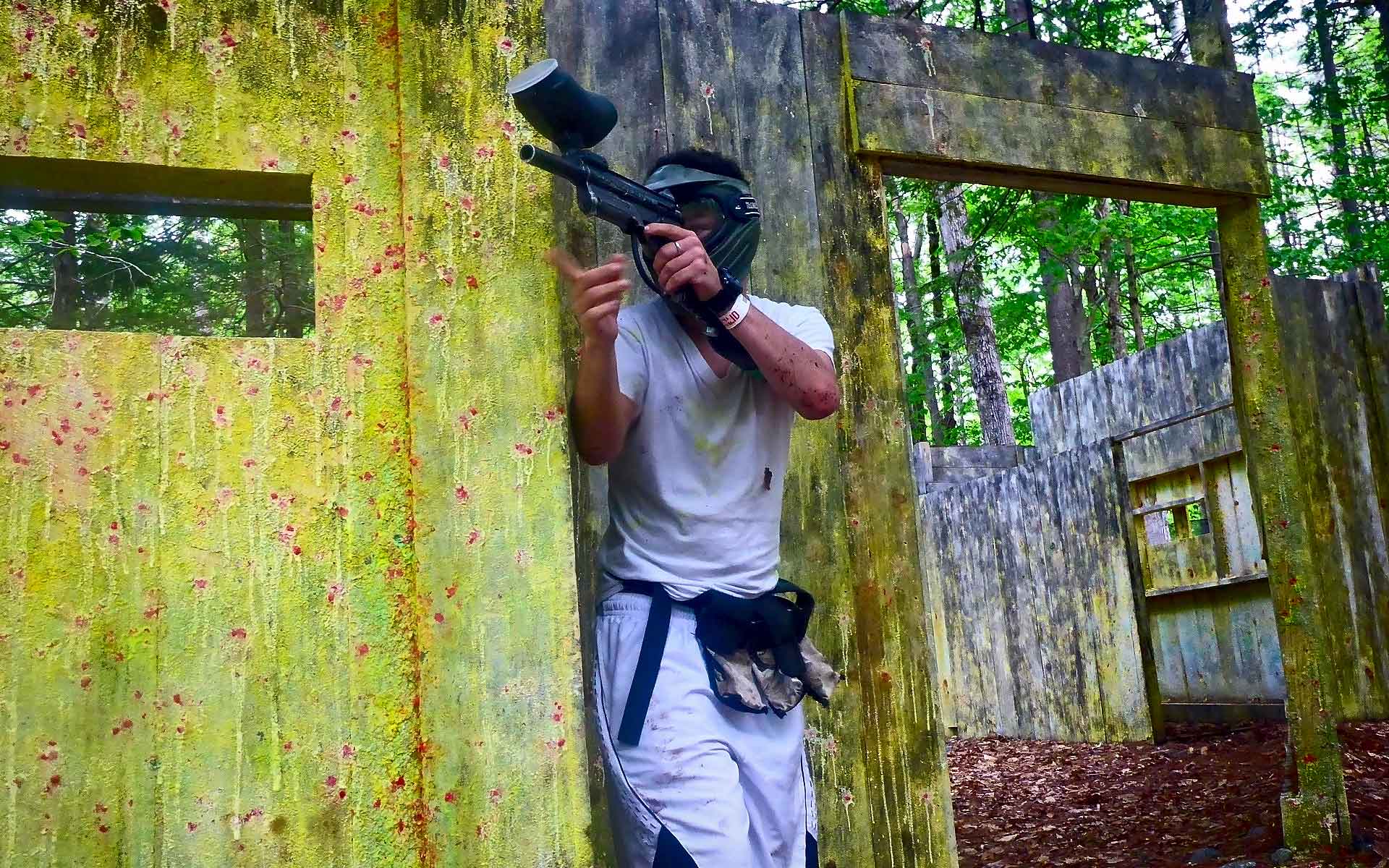 Person in white paintball gear aims a gun from behind a concrete barrier. Green trees in background.