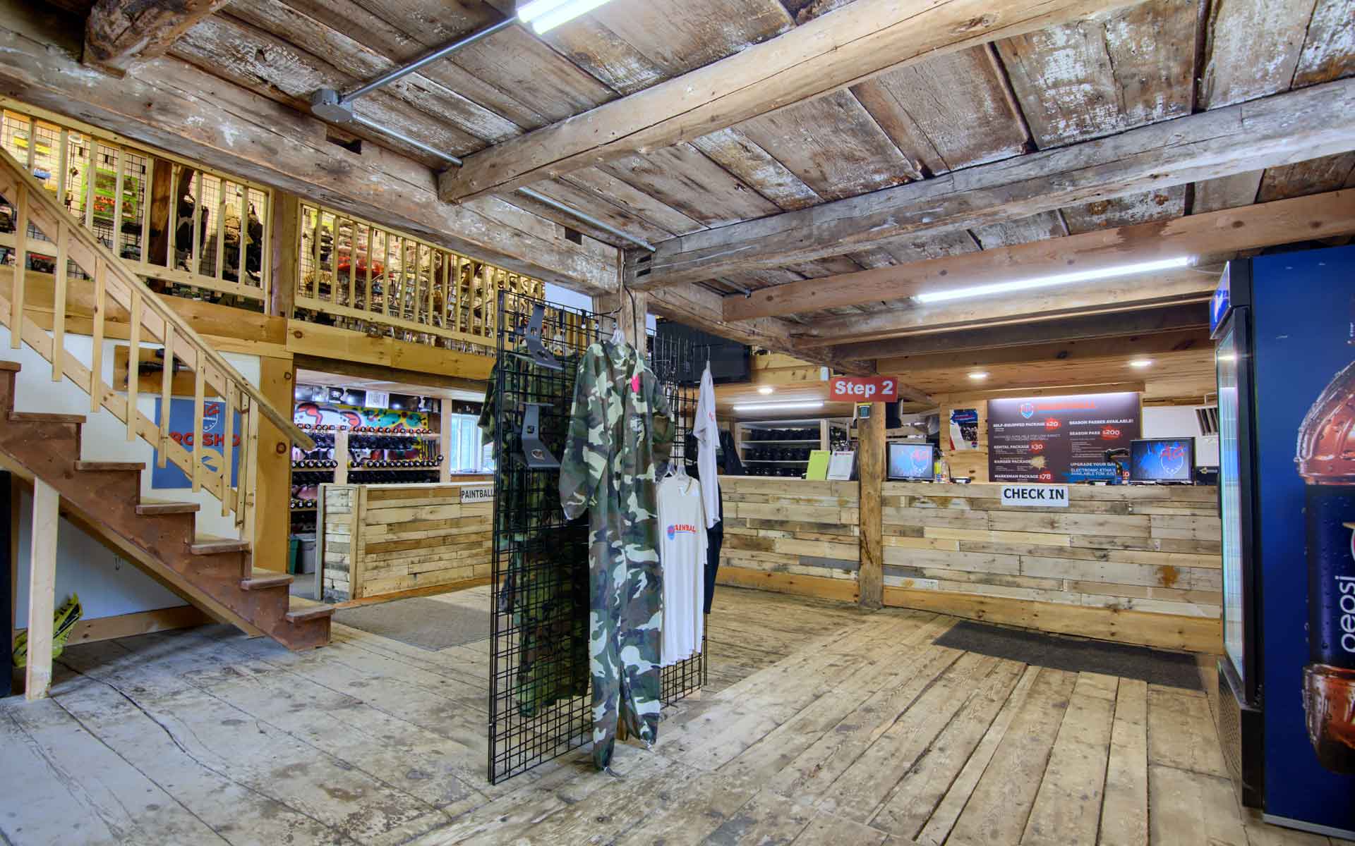 Inside a rustic store, clothing racks with camouflage suits, wooden staircase and countertops.
