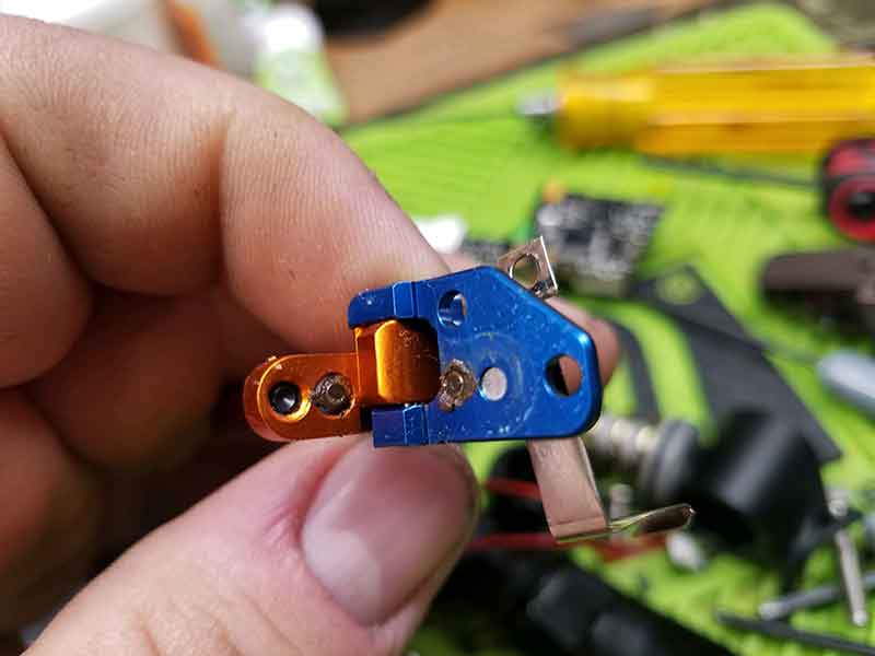 Hand holding a blue and orange metal trigger assembly. Close-up shows small holes and metal components.