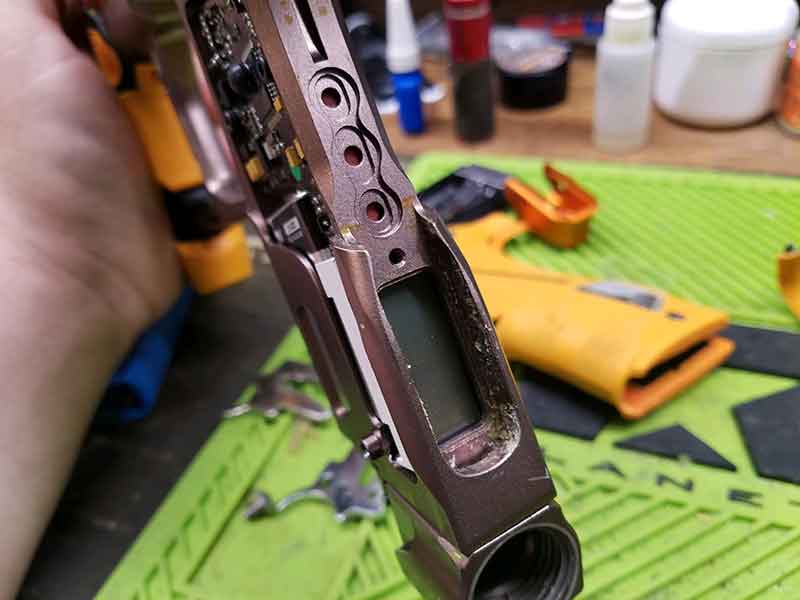 Disassembled paintball gun body, showing internal components and screen, with yellow and metallic parts, on a workbench.