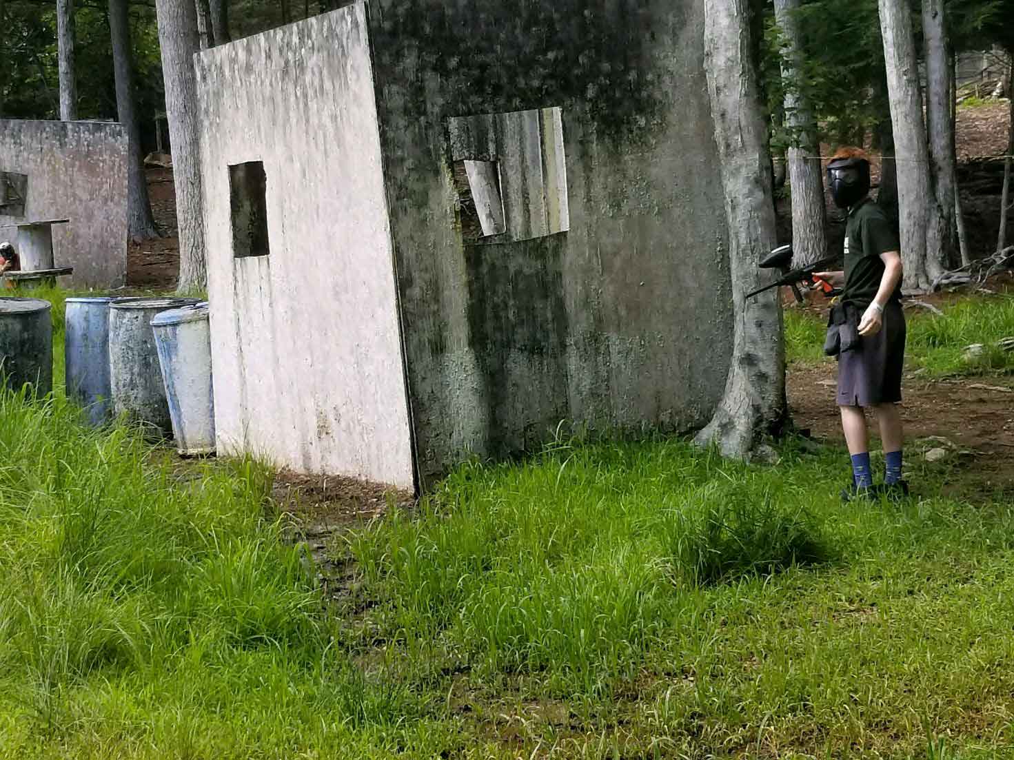 Person with paintball gear aiming a marker near a concrete structure in a wooded area.