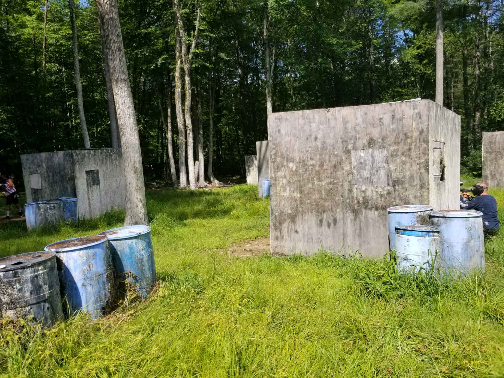 Paintball field with concrete bunkers and blue barrels amidst trees; players visible.