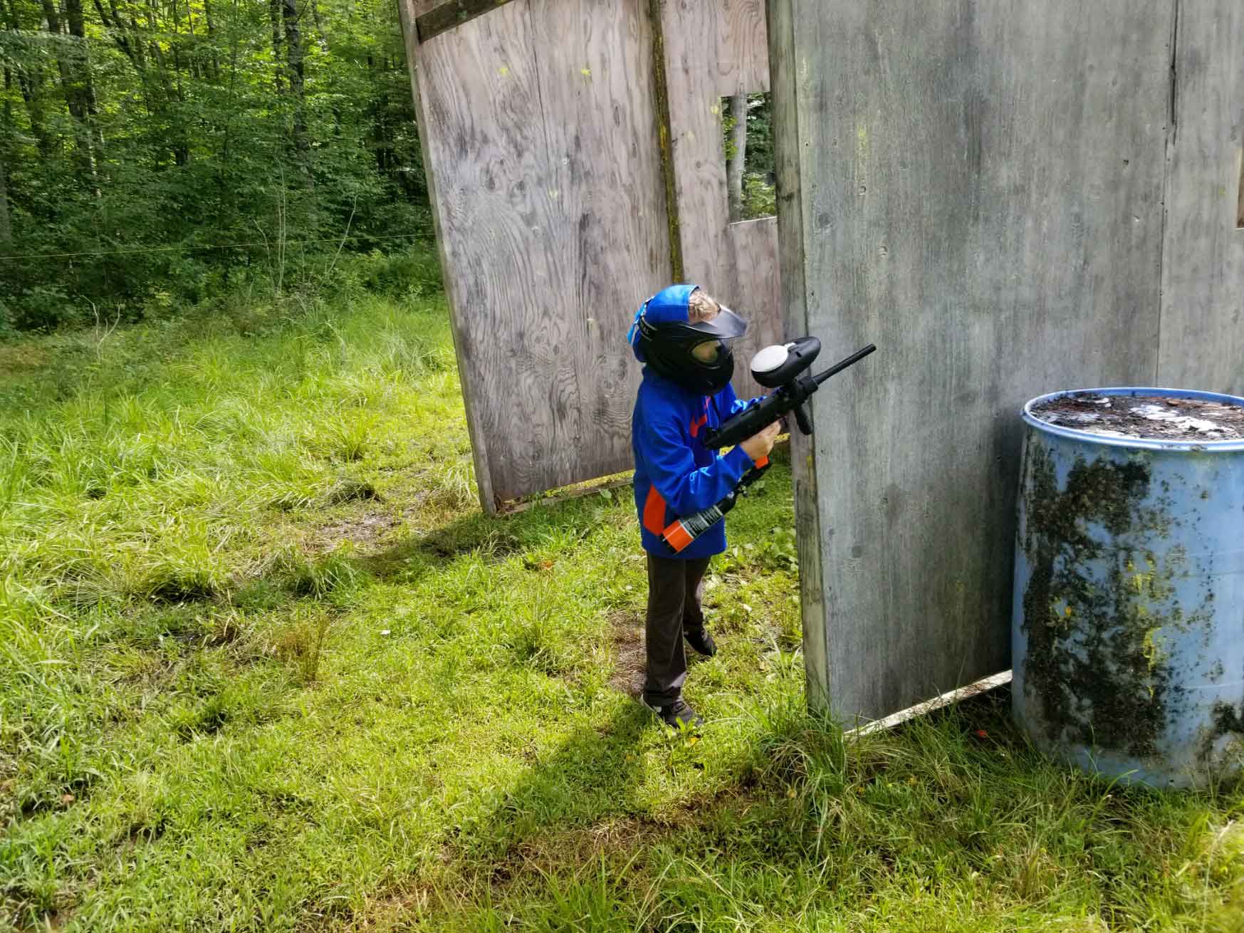 Child in protective gear holding paintball gun, playing behind wooden shelter outdoors.