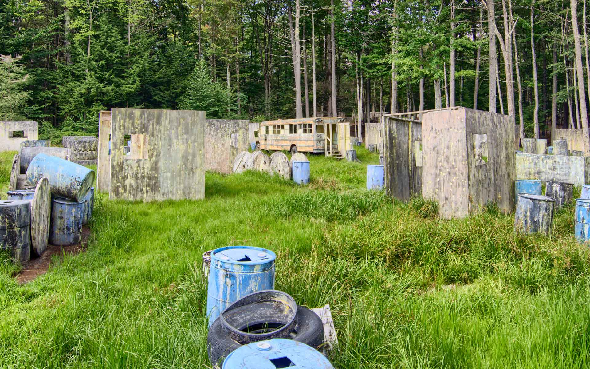 Paintball field with concrete bunkers, blue barrels, tires, and a bus in a grassy, wooded area.