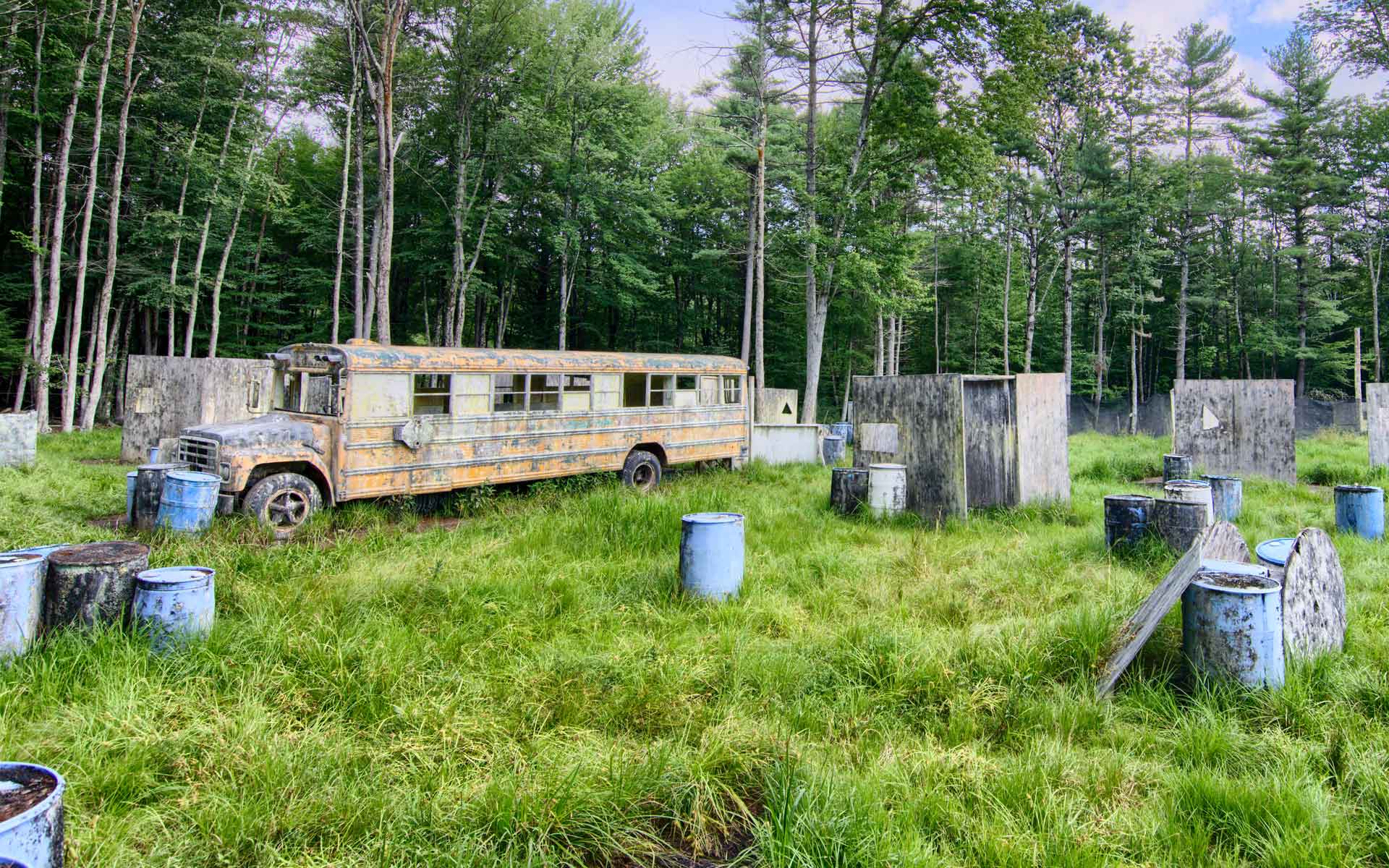 A paintball field with a weathered bus, wooden bunkers, and blue barrels amidst lush green grass.