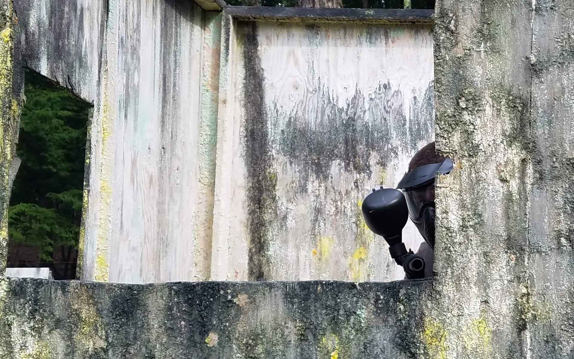 A person with a paintball gun peeks out from a weathered concrete structure at a paintball field.