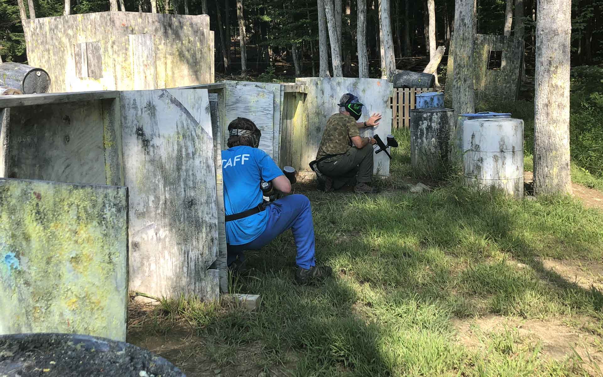 Two people playing paintball, one crouched behind a wall, another firing. Green grass, forest background.