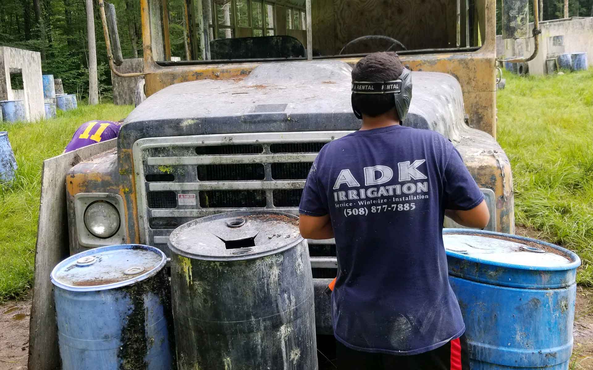 Person in paintball gear stands behind barrels, near a dilapidated school bus, at an outdoor paintball field.