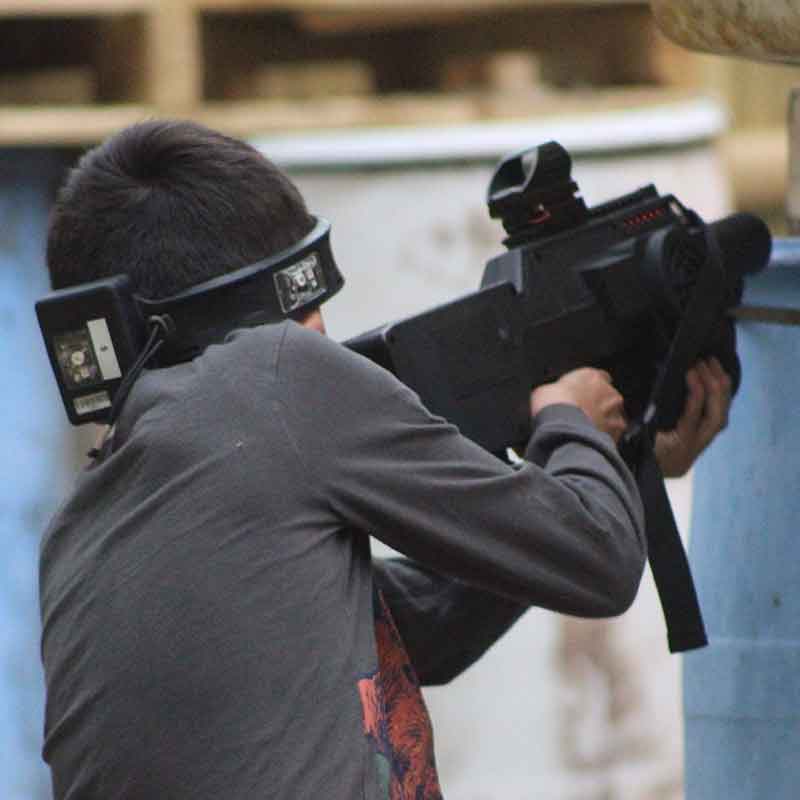 Boy wearing a laser tag headgear aims a black laser gun in an outdoor arena.