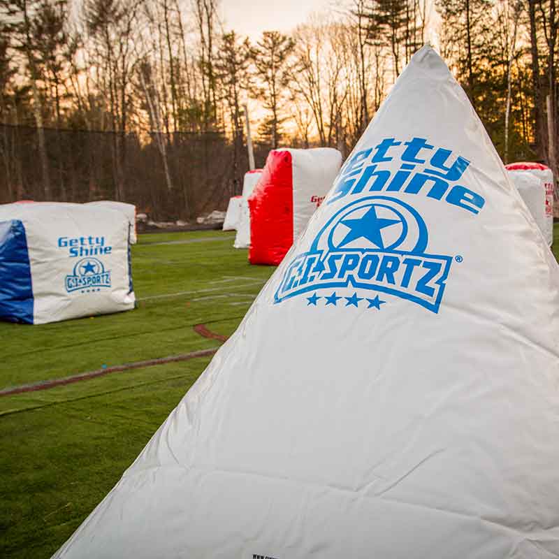 White and blue inflatable paintball bunkers on a turf field, with trees in the background.
