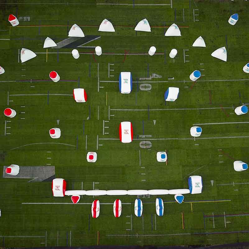 Aerial view of a paintball field; white, red, and blue inflatable bunkers on green grass.