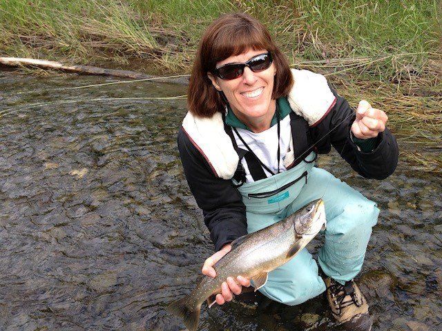 A smiling person in fishing gear holds a freshly caught fish while kneeling in a shallow, rocky river.