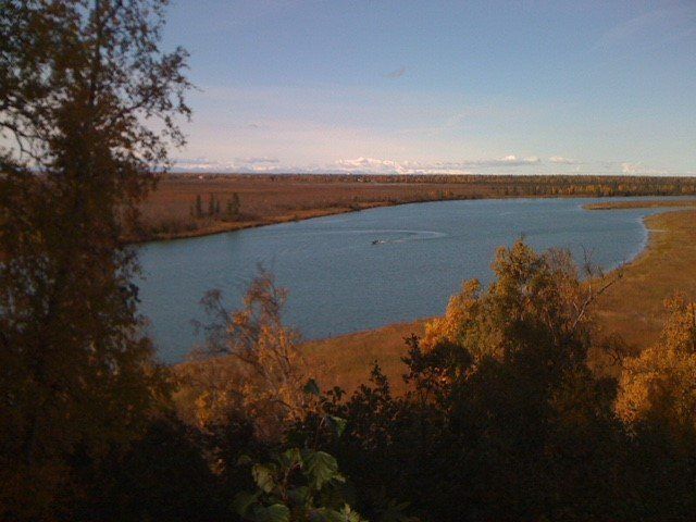 A scenic view of a river winding through a brown, autumnal landscape under a blue sky, framed by golden-leafed trees.