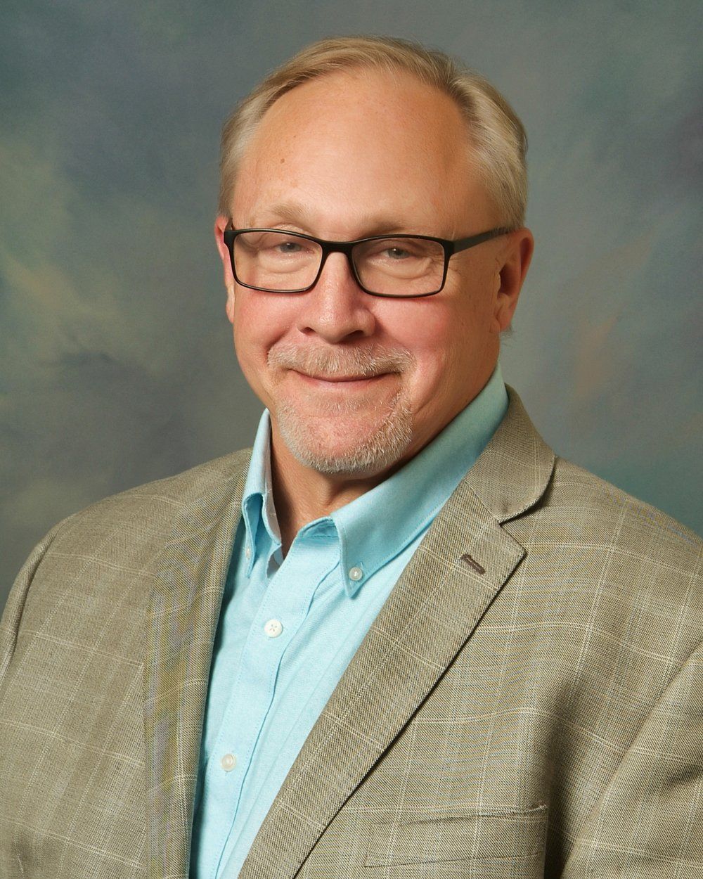 A man with glasses and a goatee, wearing a light blue collared shirt under a gray blazer, smiles against a gray background.