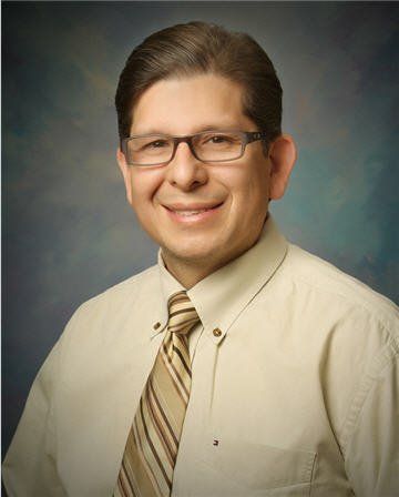 A smiling person wearing glasses, a button-down shirt, and a striped tie against a blue backdrop.