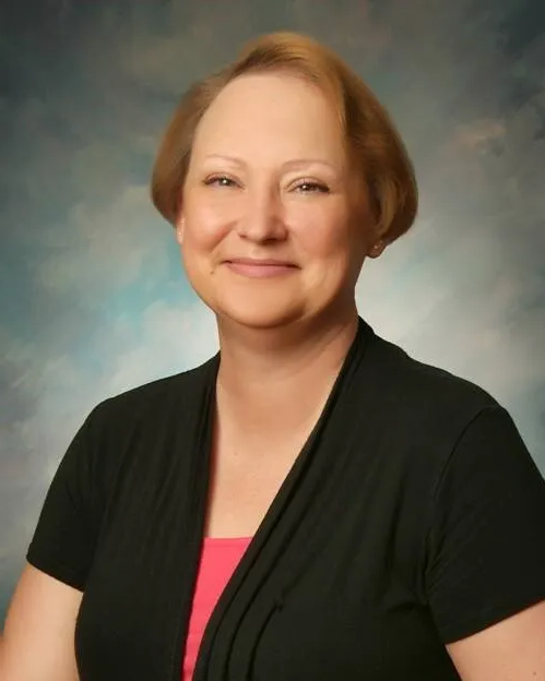 Headshot of a person with light brown hair wearing a black top over a bright pink shirt against a blue-grey background.
