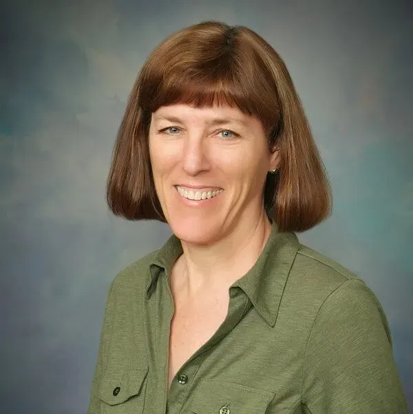 A woman with shoulder-length brown hair and bangs smiles while wearing a green collared shirt against a blue backdrop.