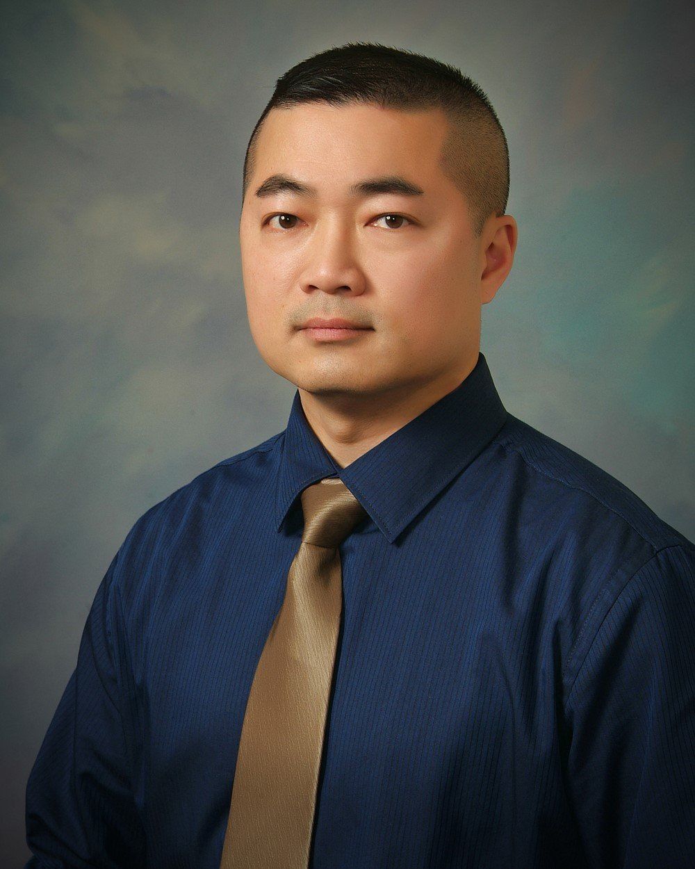 A person wearing a dark blue button-down shirt and tan tie against a textured, light-to-dark blue gradient background.