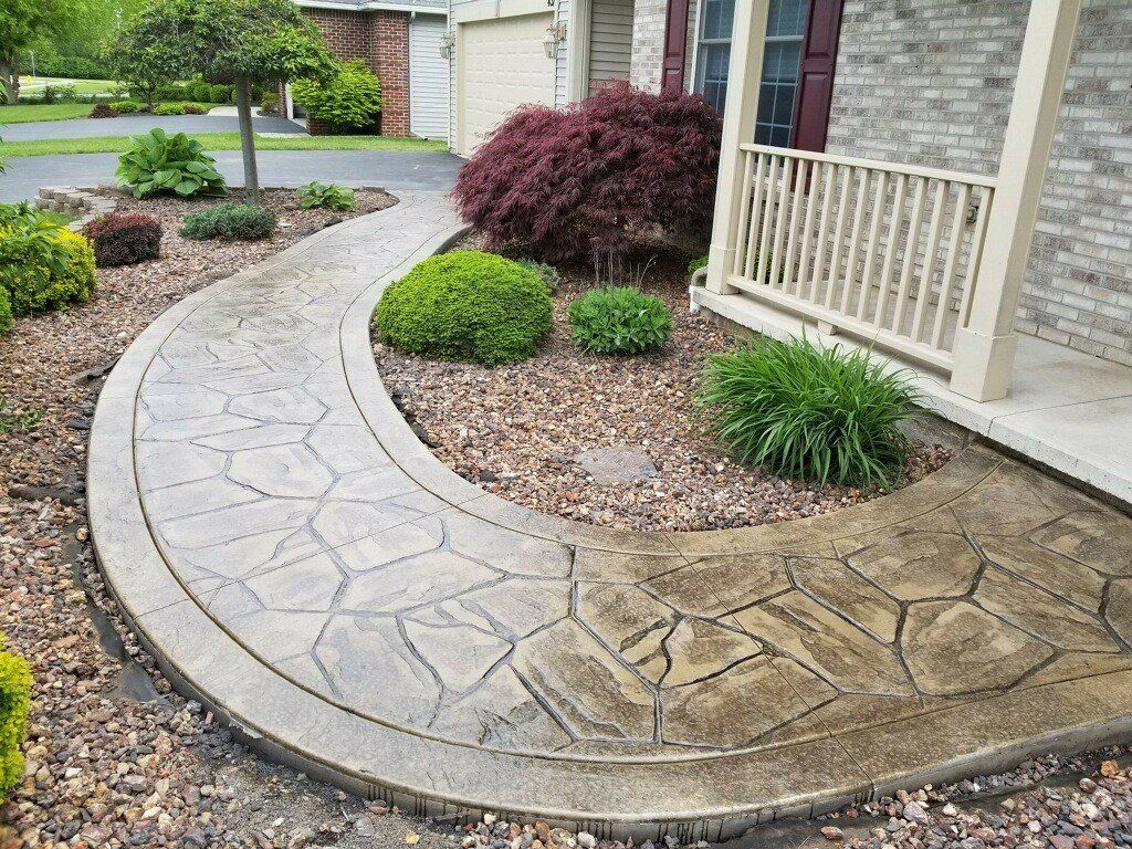 Curving stamped concrete walkway leading to a house porch, surrounded by landscaping and bushes.