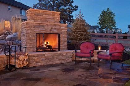 Outdoor stone fireplace with burning fire, red chairs, and stacked firewood.