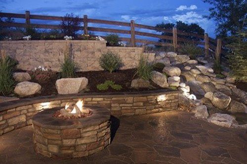 Outdoor stone fire pit on a patio, with a tiered stone wall and wooden fence in the background at dusk.