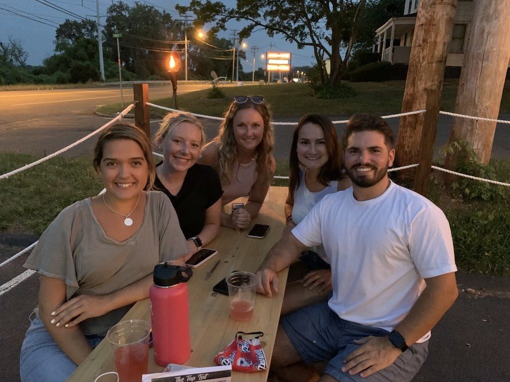 Group of five people at an outdoor table. Drinks and a water bottle are on the table. Evening setting.