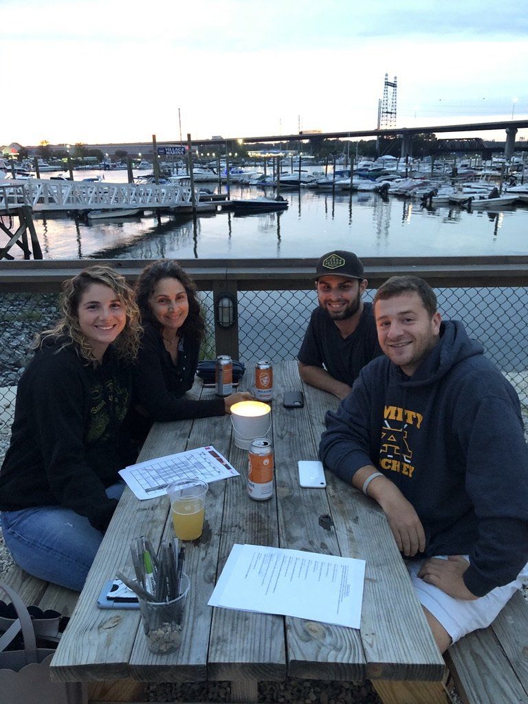 Four people seated at a waterfront table, drinks, marina background, dusk.