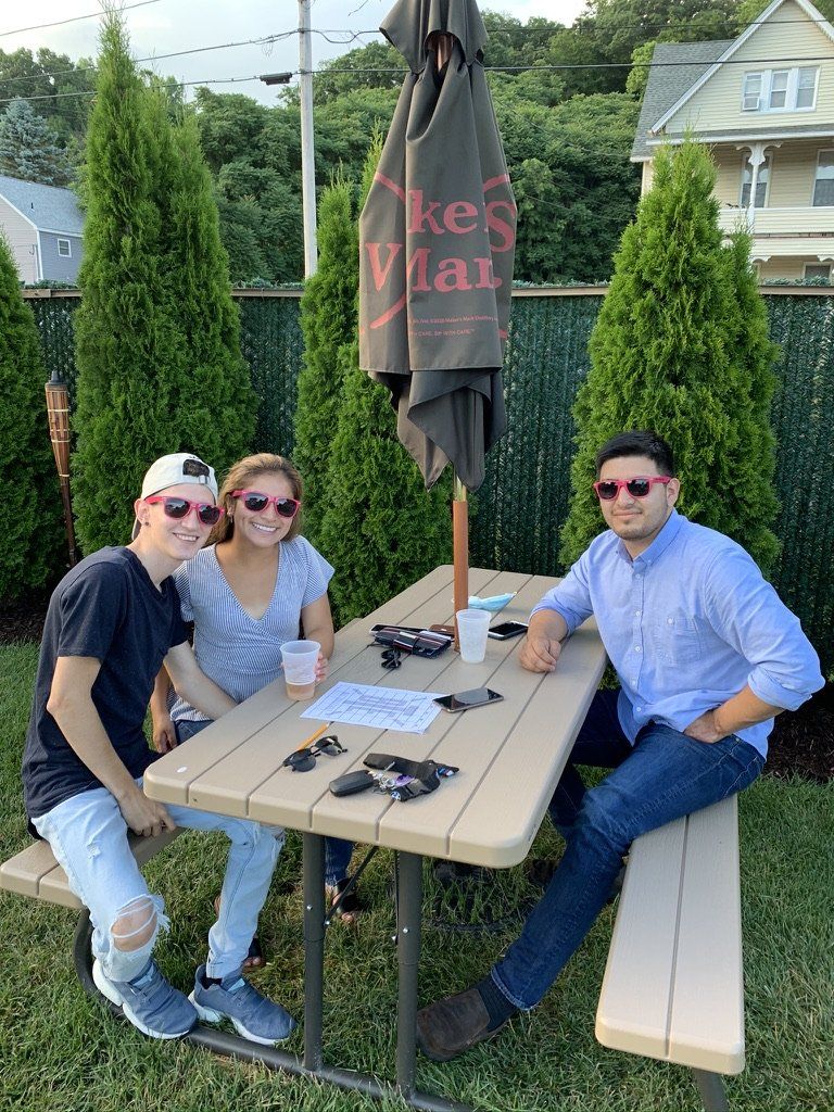Three people wearing pink sunglasses sit at a picnic table, an umbrella overhead, outdoors.