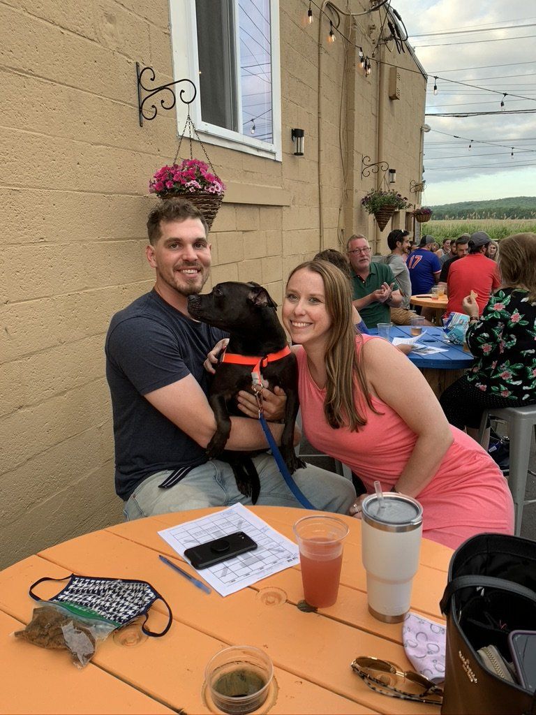 Couple at outdoor brewery with a black dog. Orange table, beers, and a mask. Other people in the background.