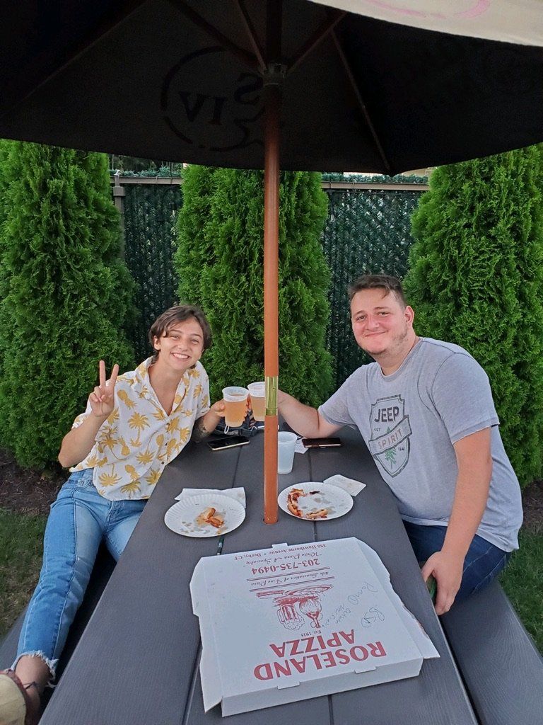 Two people under an umbrella at a picnic table, eating pizza and drinking. One gives a peace sign.