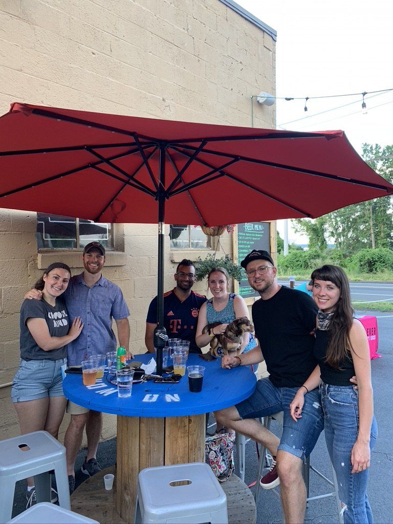 Group of people at outdoor bar under red umbrella, smiling and holding drinks.