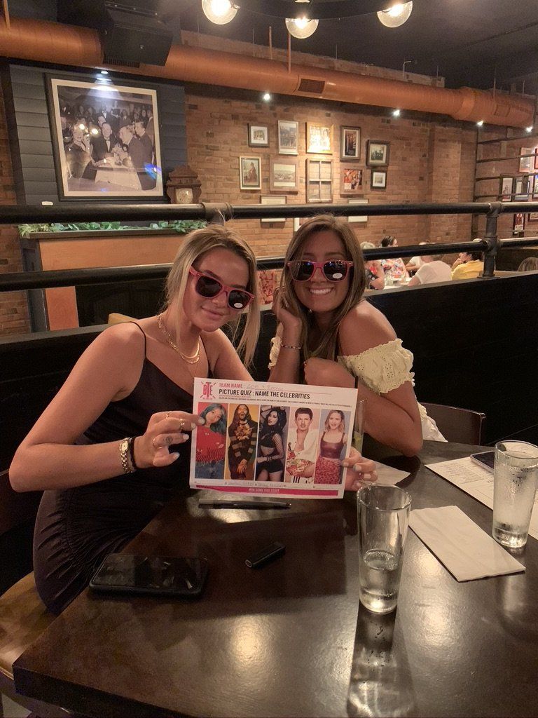 Two women in sunglasses at a restaurant holding a flyer with images of people.
