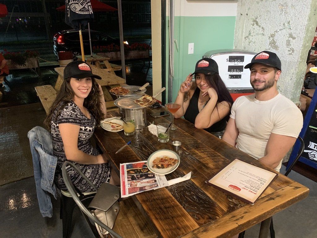 Three people wearing hats sit at a wooden table in a restaurant. Pizza and drinks are visible.