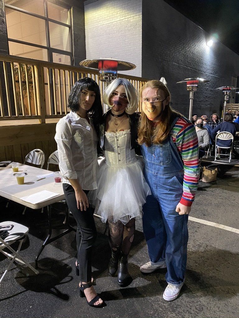 Three people in Halloween costumes outdoors: a vampire, a bride, and Chucky.
