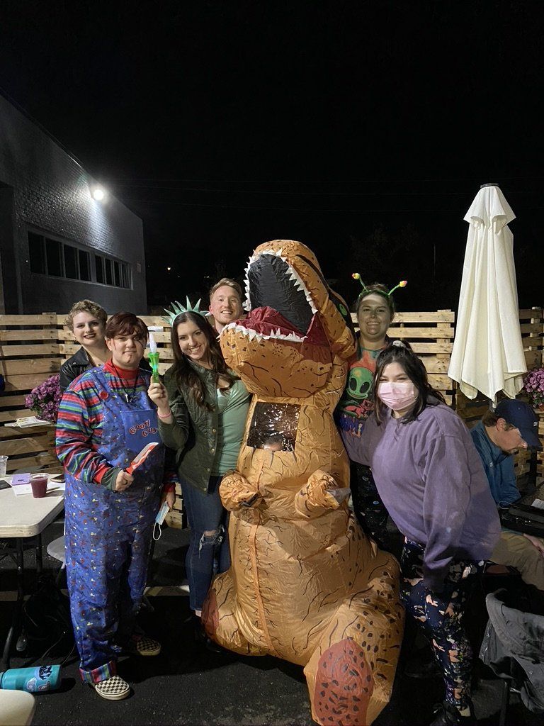 Group of people in Halloween costumes posing with an inflatable dinosaur outdoors at night.